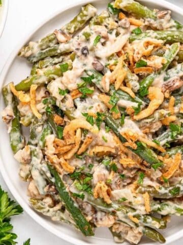 A plate of green bean casserole topped with fried onions, accompanied by a bowl of mashed potatoes. Fresh parsley and mushrooms are nearby on a white surface.