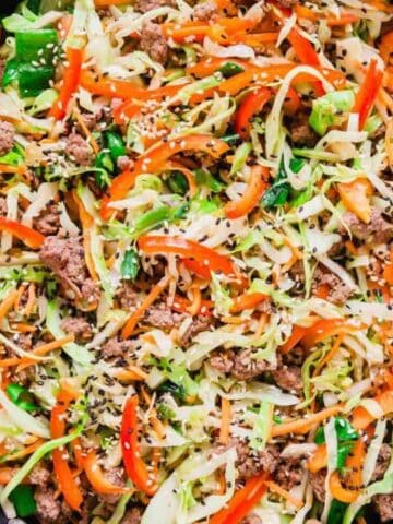 Pan of stir-fried vegetables and beef, including cabbage, carrots, red peppers, and topped with sesame seeds, on a striped cloth.