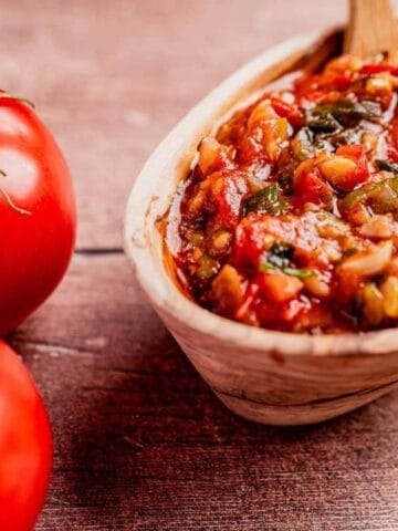 Wooden bowl of salsa with tomatoes, peppers, and onions, next to two tomatoes and a green onion on a wooden surface.