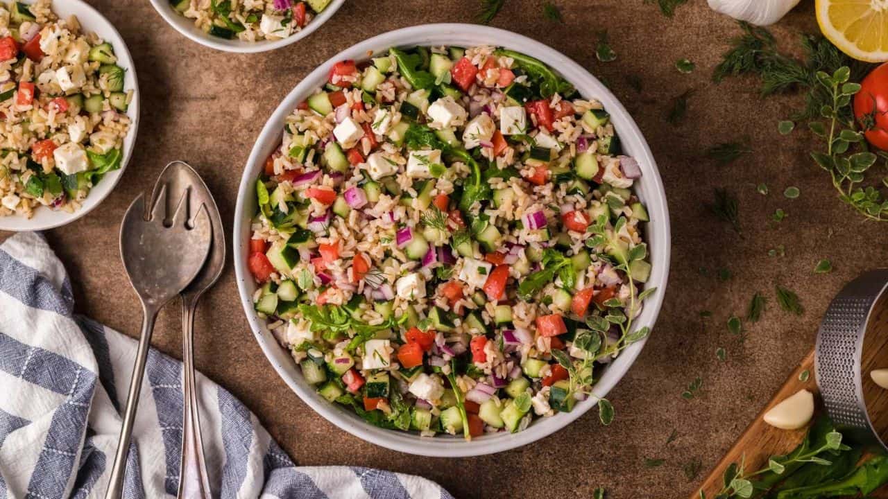 A large bowl of mixed salad with chopped cucumbers, tomatoes, red onions, feta, and herbs on a wooden surface. A blue and white cloth, salad servers, and herbs are nearby.