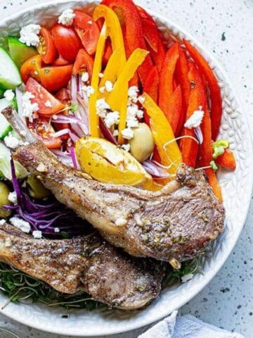 A bowl with grilled lamb chops, sliced cucumbers, red and yellow peppers, tomatoes, red onions, olives, and feta cheese on a speckled countertop. Seasoning and garlic visible nearby.