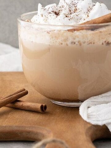 A clear glass cup of frothy coffee topped with whipped cream and sprinkled cinnamon, set on a wooden board with two cinnamon sticks. A white cloth is draped beside it.
