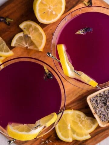 Two purple-hued cocktails garnished with lemon slices and lavender, served on a wooden board with scattered lemon slices, dried lavender, and a yellow cloth nearby.
