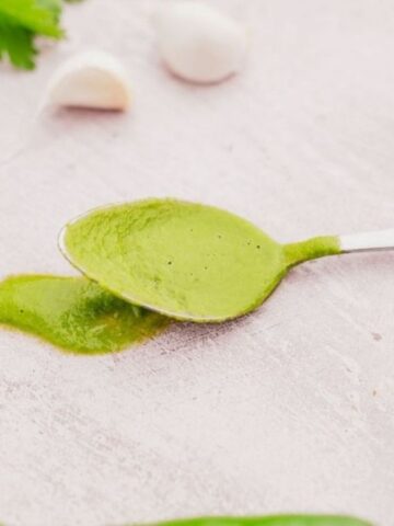 A spoon with green sauce on a light background, surrounded by fresh green herbs, garlic cloves, and a green chili pepper.