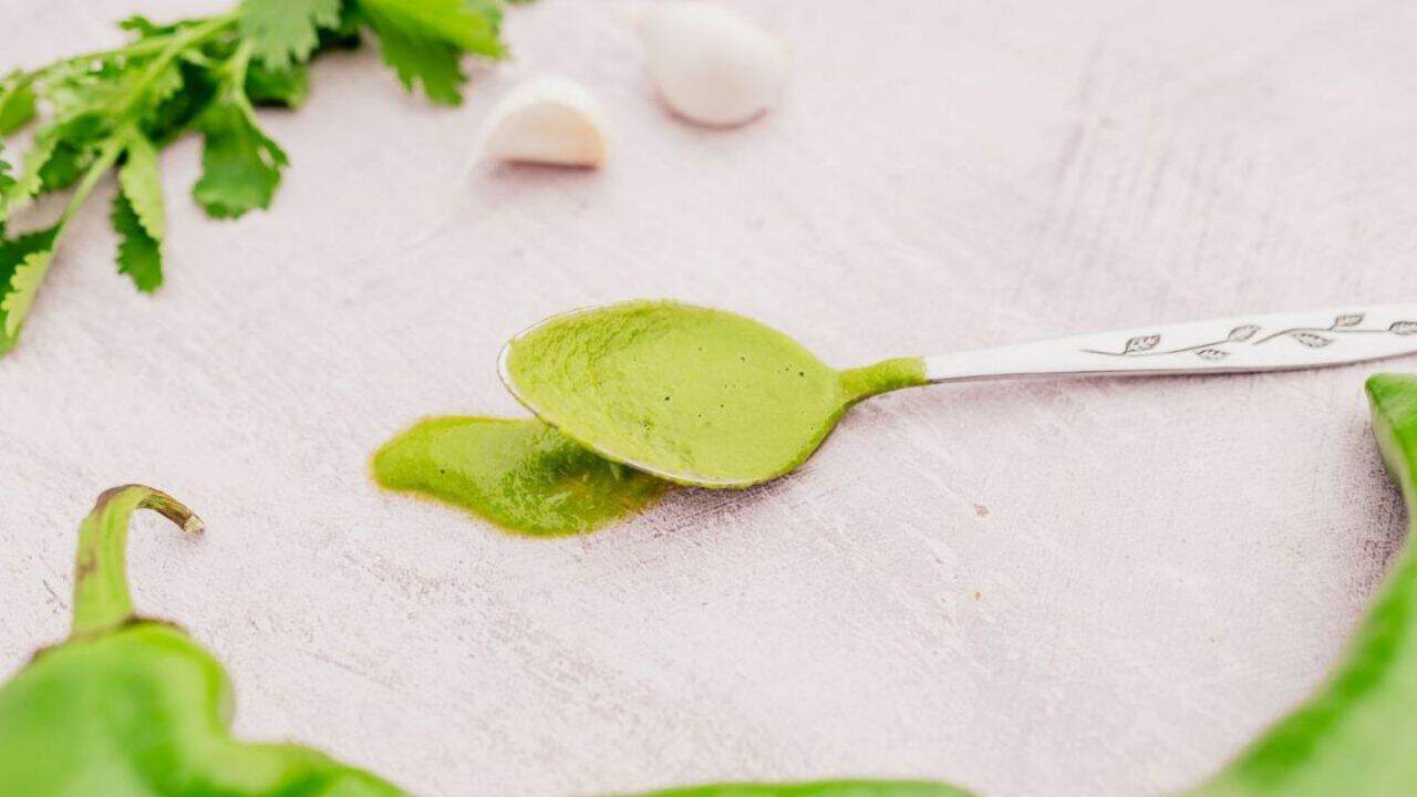 A spoon with green sauce on a light background, surrounded by fresh green herbs, garlic cloves, and a green chili pepper.