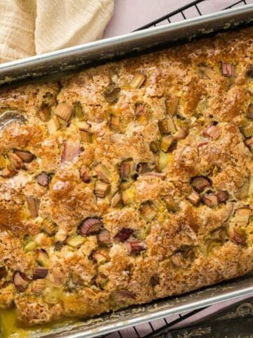 A freshly baked rhubarb cake in a rectangular pan on a cooling rack, with a cloth, lemon, and rhubarb stalks nearby.