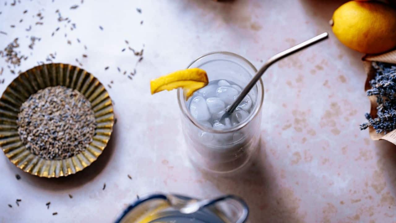 Glass of iced lavender lemonade with a metal straw and lemon wedge, surrounded by dried lavender, lemons, and a yellow dish on a light-colored surface.