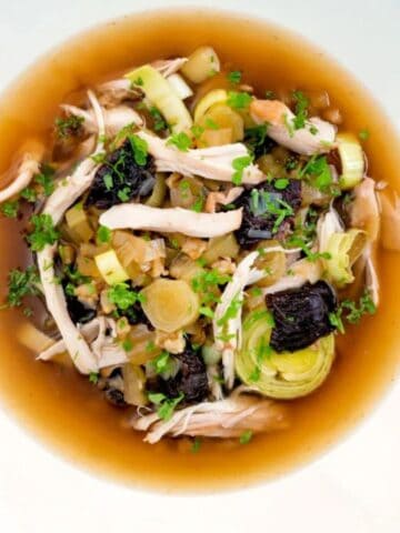 A bowl of broth with shredded chicken, sliced leeks, prunes, and herbs, served on a wooden table with a cloth and slices of bread.
