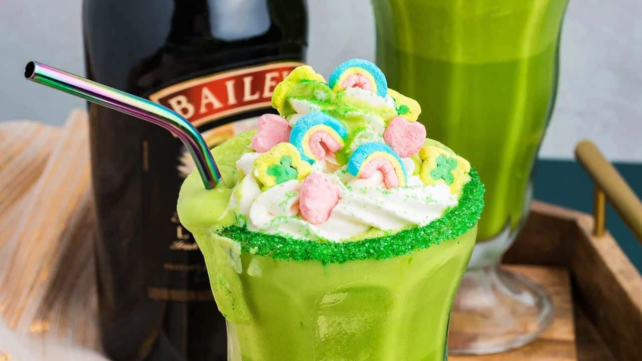 A green milkshake topped with whipped cream, colorful marshmallows, and a green sugar rim, next to a Baileys bottle.
