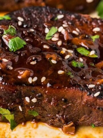 A cooked steak glazed with a dark sauce, garnished with sesame seeds and green herbs, on a white plate.