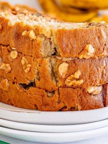 Slices of nut-studded banana bread stacked on a white plate, with bananas and a bag in the background.