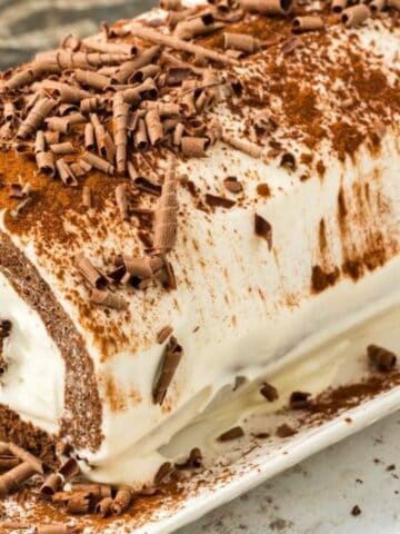Chocolate roll cake topped with chocolate shavings and icing, displayed on a white plate.