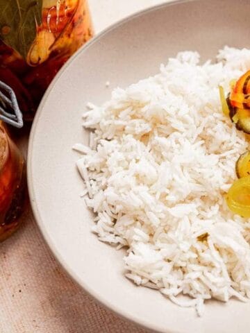 A bowl of white rice with pickled vegetables including carrots and peppers. Two jars of pickled vegetables beside the bowl. A spoon is placed in the bowl.