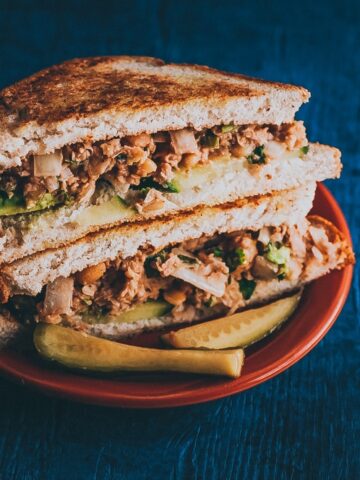 A sandwich with two stacked halves filled with meat, lettuce, and onion on whole-grain bread, served with pickle slices on a red plate against a dark blue background.