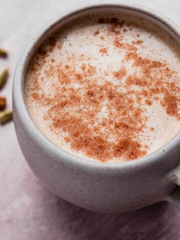 A mug of frothy coffee sprinkled with cinnamon. Nearby are cinnamon sticks, coffee beans, and cardamom pods on a textured surface.