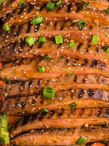 Grilled steak slices on a bed of broccoli and carrots, garnished with sesame seeds and chopped green onions in a Staub dish.