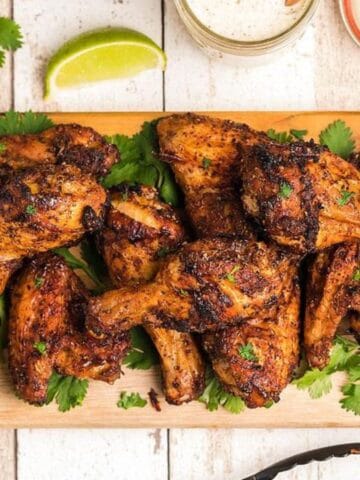 A wooden board topped with seasoned, grilled chicken wings garnished with cilantro. A small bowl of dipping sauce is on the side, along with lime wedges and sauces.
