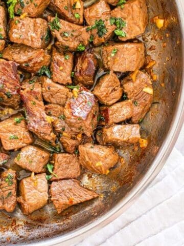 Cooked steak bites garnished with herbs in a skillet, with garlic cloves and a wooden spoon nearby on a marble surface.