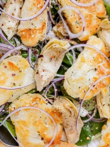 Salad with cooked halloumi, red onion rings, artichoke hearts, and greens, served in a white bowl with two metal serving spoons.