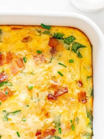 Baked casserole with eggs, bacon, and spinach in a white dish. Fresh spinach leaves in a bowl beside it.