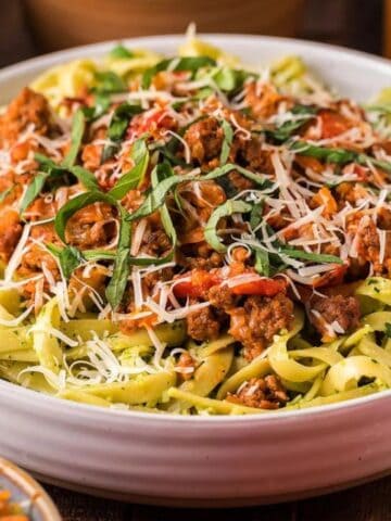 A bowl of fettuccine pasta topped with tomato sauce, ground meat, shredded cheese, and fresh basil, surrounded by a wooden table setting.