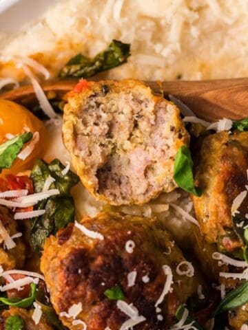 A plate of meatballs atop creamy polenta, garnished with grated cheese, fresh greens, and cherry tomatoes. A wooden spoon rests on the dish.