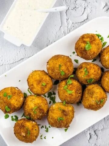 A white rectangular plate with twelve golden-brown fried balls garnished with herbs, accompanied by a small dish of creamy white dipping sauce.