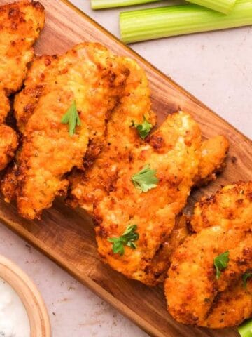 Four pieces of fried chicken tenders are arranged on a wooden board with celery sticks, a bowl of buffalo sauce, and a bowl of ranch or blue cheese dip.