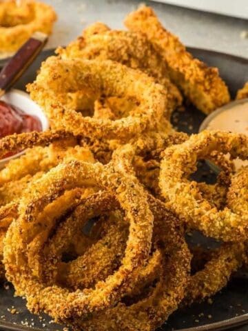 A black tray filled with crispy onion rings, served with small bowls of ketchup and a creamy dipping sauce, and two dipping forks.