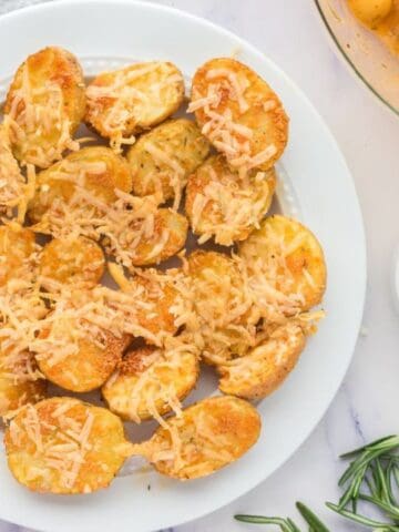 A white plate with roasted potato halves topped with shredded cheese, next to bowls of sliced potatoes, spices, and fresh rosemary on a marble surface.