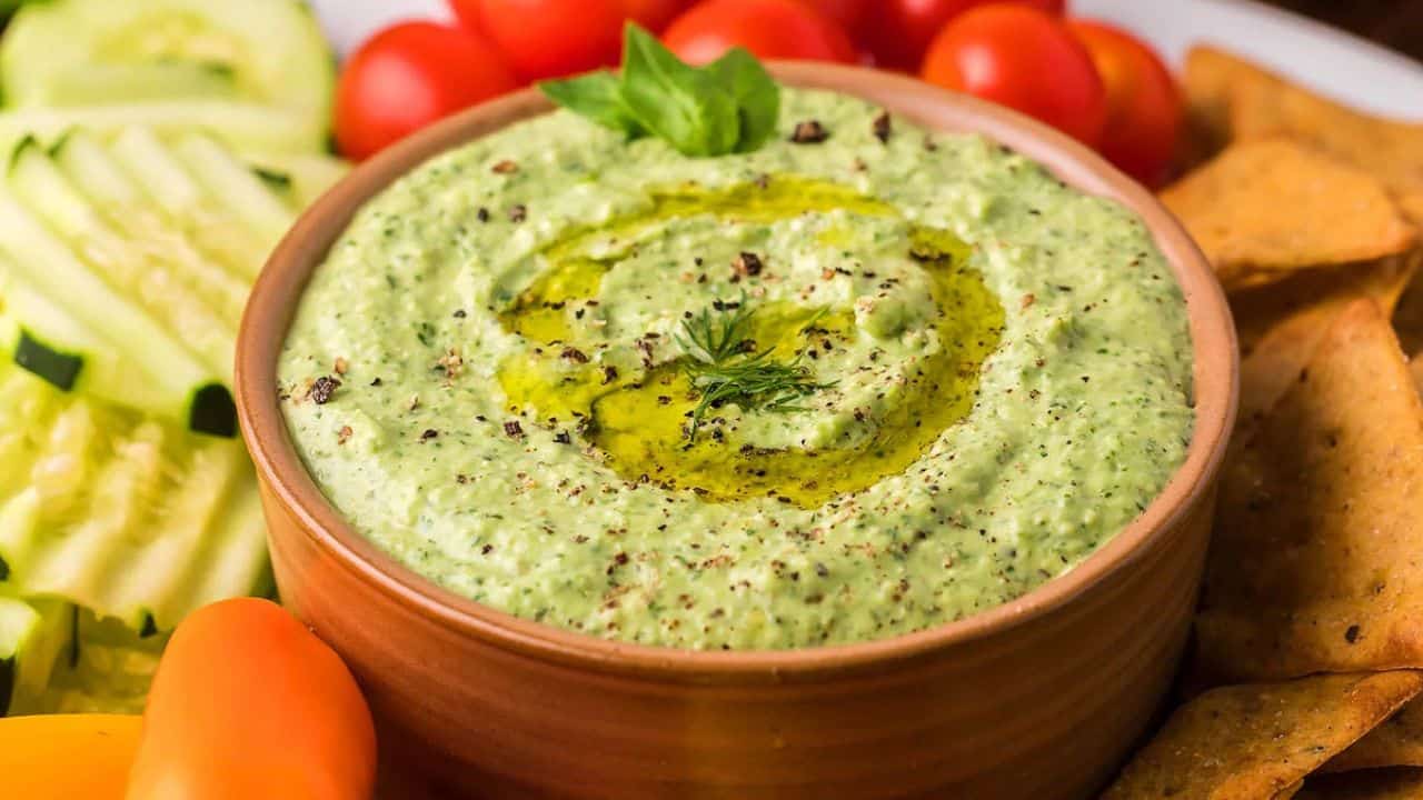 A bowl of green dip garnished with herbs and olive oil, surrounded by sliced cucumbers, cherry tomatoes, and chips.