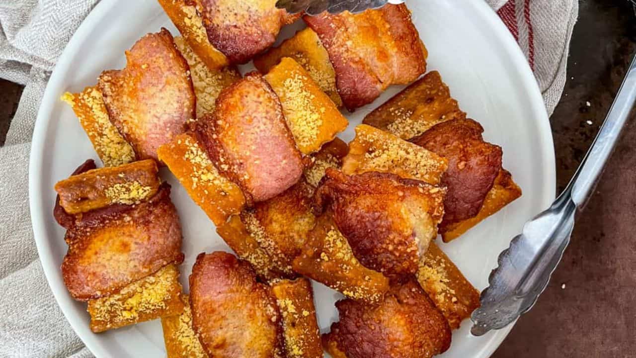 Plate of crispy bacon-wrapped breadsticks, topped with a sprinkle of cheese, with tongs on the side.