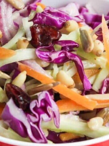 A colorful salad in a red bowl with purple cabbage, carrots, celery, pine nuts, pumpkin seeds, and dried cranberries.