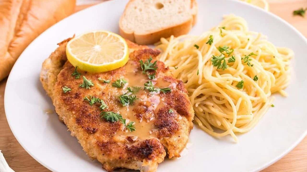 A plate with breaded chicken topped with sauce and a lemon slice, served with a side of spaghetti garnished with parsley and a slice of bread in the background.