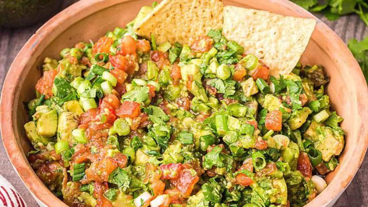 A bowl of chunky guacamole with diced tomatoes, green onions, and cilantro, garnished with tortilla chips.