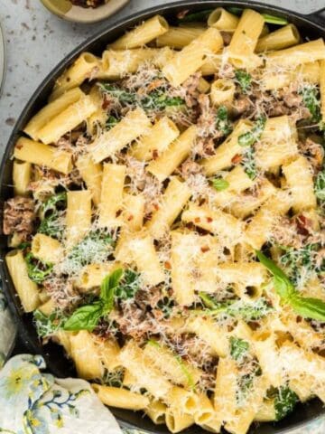 A skillet of cooked rigatoni pasta with ground meat, herbs, and grated cheese. Nearby are plates, forks, a wooden spoon, and a block of cheese with a grater.