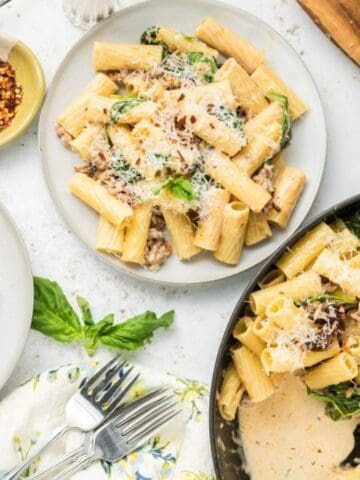 Three plates of rigatoni pasta with sauce, spinach, and grated cheese are served alongside a pan with the same dish, red pepper flakes, and sliced bread on a white surface.