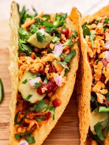Three hard-shell tacos with avocado, cilantro, onions, and sauce on a wooden board, served with lime slices and a bowl of red salsa.