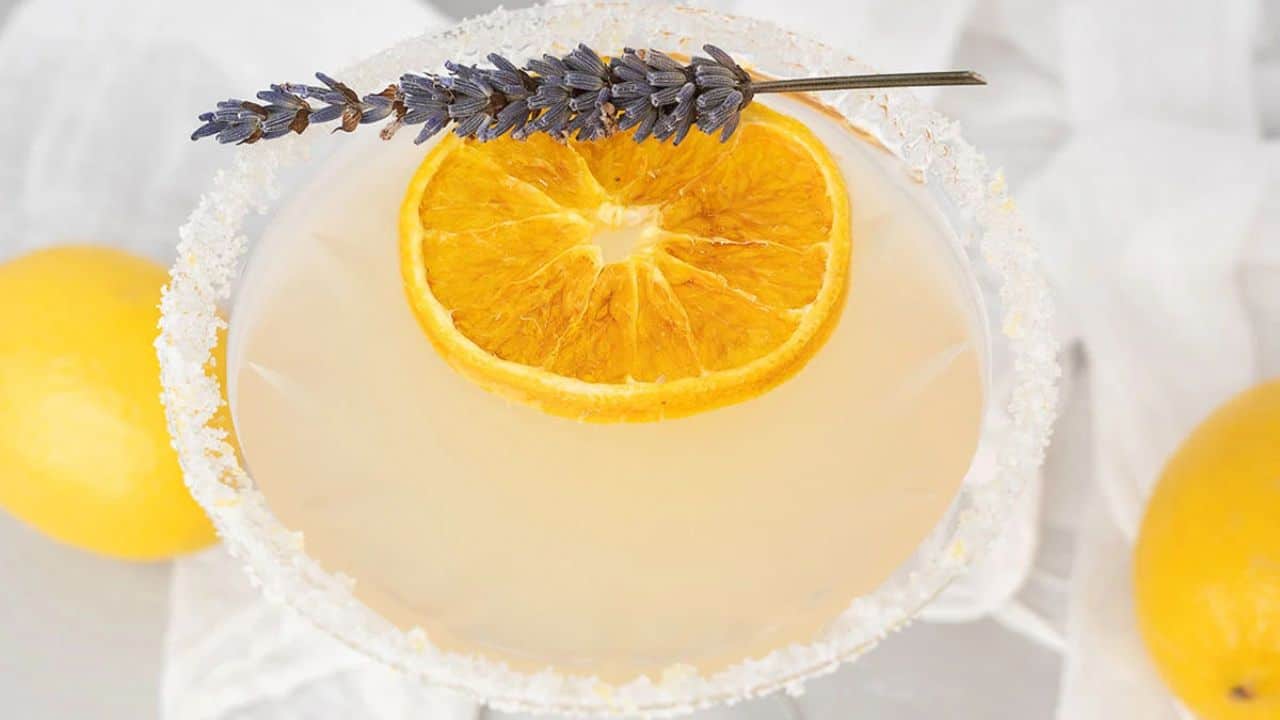 A cocktail garnished with a dried orange slice and a sprig of lavender, served in a glass with a sugared rim; lemons are visible in the background.