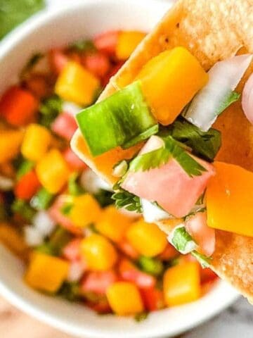 A hand holds a tortilla chip topped with mango salsa, with a bowl of salsa and fresh cilantro in the background.