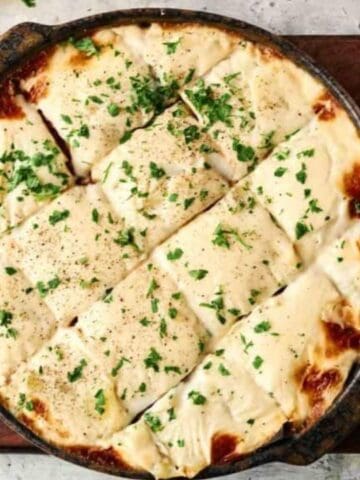 A baked dish of lasagna in a round pan, topped with melted cheese and chopped parsley, placed on a wooden board with a small bowl of herbs and a white pepper grinder nearby.