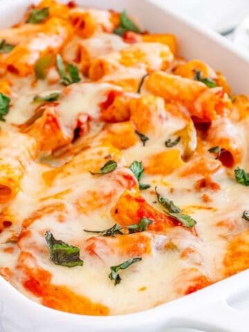 A white casserole dish filled with baked rigatoni pasta, topped with melted cheese and garnished with basil, sits on a table next to a bowl of shredded cheese.
