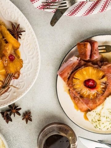 A plate with glazed ham topped with pineapple and cherry, mashed potatoes with herbs, and mixed vegetables; a larger serving of ham is on a platter nearby.