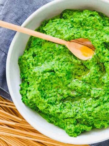 A white bowl filled with green pesto sauce and a wooden spoon, placed on a gray napkin next to uncooked spaghetti on a wooden board.
