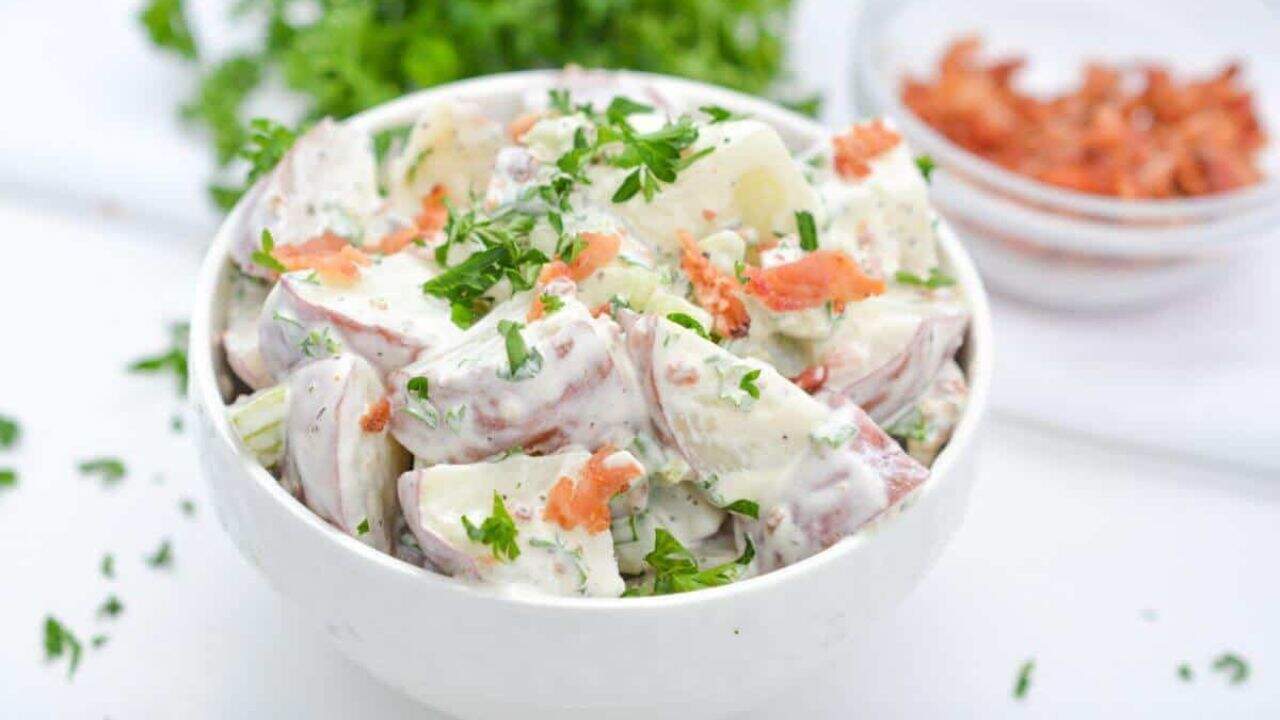 Bowl of potato salad garnished with fresh herbs and bacon on a white background.