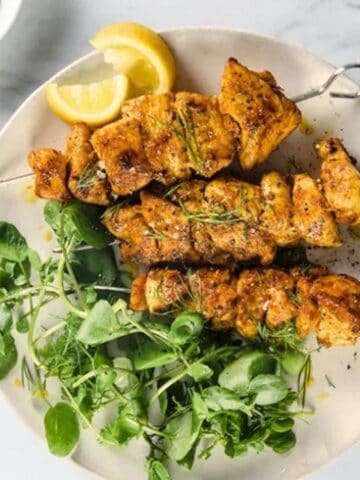 Three grilled chicken skewers on a plate with fresh greens, lemon wedges, and a bowl of sauce on the side.