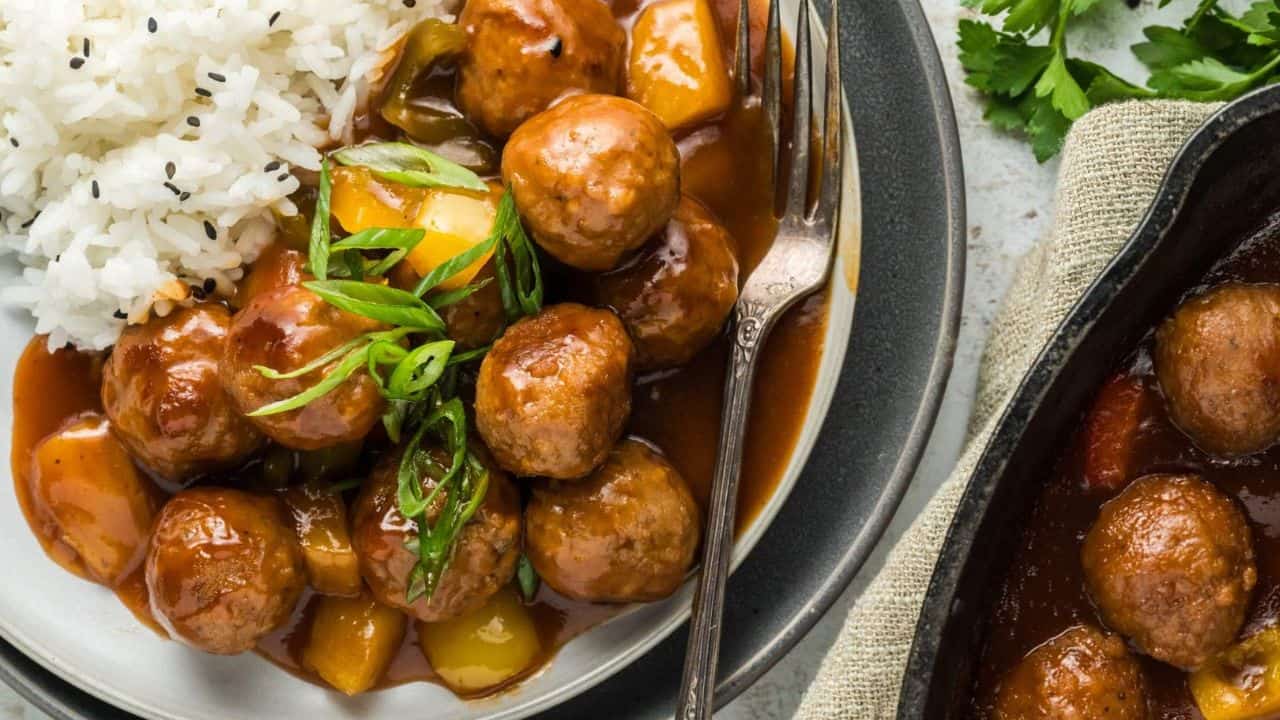 Plate of rice and meatballs in a brown sauce with vegetables, garnished with sliced green onions; fork on the plate, with a skillet nearby.