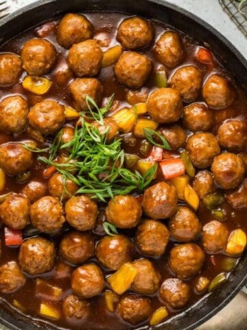 A skillet filled with meatballs in sauce, topped with greens, alongside pineapple slices and a bowl of white rice.