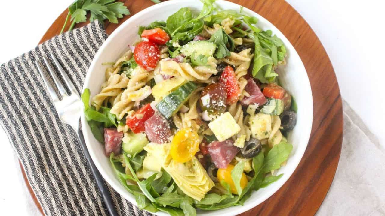 A bowl with spiral pasta, vegetables, pepperoni, and arugula.