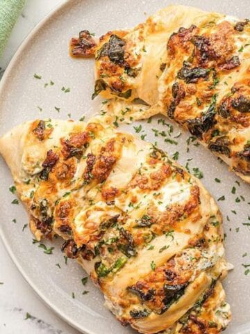 Two stuffed chicken breasts topped with herbs are served on a white plate, with a green cloth and a bowl of red sauce nearby.